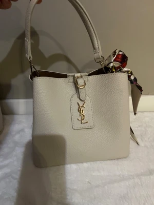 Vit YSL handväska med scarfdetalj - Stilren vit handväska från YSL i strukturerat läder med guldfärgad logga framtill. Väskan har ett kort handtag och en färgglad scarf knuten på sidan som extra detalj. Klassisk fyrkantig form och elegant design som lyfter vilken outfit som helst.