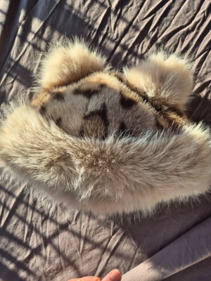 Fuskpälsmössa med öron - Säljer en fluffig mössa i fuskpäls med söta öron på toppen. Mössan är beige med mörkare fläckar och har en bred, mjuk kant runt om. Perfekt för dig som vill sticka ut och hålla dig varm med en lekfull stil.