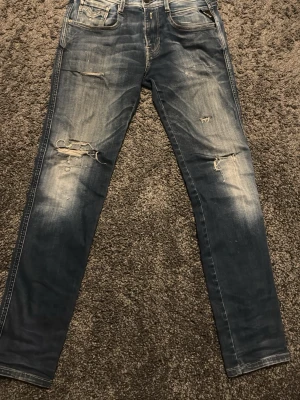 Replay Anbass jeans med egengjorda slitningar - Säljer ett par blå skinny jeans med slitningar och hål på framsidan. Jeansen har klassisk femficksdesign, ljusa tvättade partier och smal passform. Perfekta för en avslappnad och trendig look. Priset är inte hugget i sten! Storleken är 29/32 Något kortare i storleken!