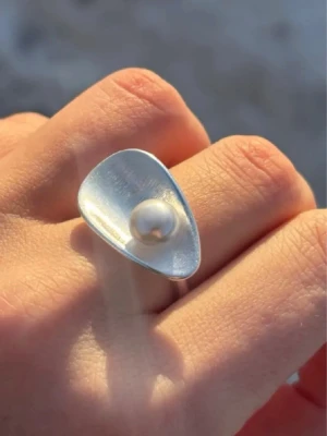 Silverring med pärldetalj - silverring med en trekantig, lätt kupad platta och en vit pärla i mitten. Ringen har en modern och stilren design som sticker ut. Tillverkad i äkta silver, vilket syns på stämplingen på insidan av ringen. Perfekt för dig som gillar statement-smycken.