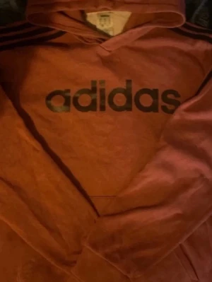 Rostbrun Adidas hoodie med huva - Säljer en snygg rostbrun hoodie från Adidas med svart logga framtill och klassiska svarta ränder på axlarna. Hoodien har huva och en stor magficka. Perfekt för dig som gillar streetwear och sportig stil.