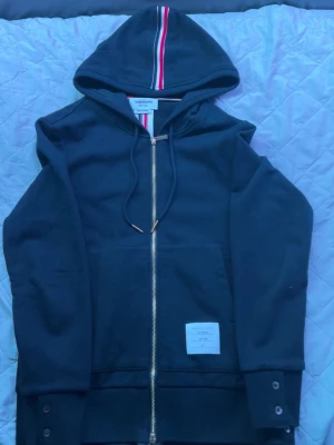 Mörk blå zip hoodie från Thom Browne - Säljer en svart hoodie från Thom Browne med dragkedja i guld, huva med snören och ribbade muddar. Tricolore-detalj i luvan och knappdetaljer vid ärmslut och nederkant. Tillverkad i mjuk bomull och har en vit patch med logga på framsidan. Det är storlek 1 vilket mot svarar Medium