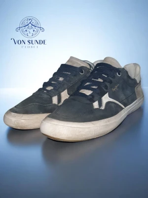 Svarta sneakers från Pepe Jeans - Snygga svarta sneakers från Pepe Jeans med vita detaljer och logga på sidan. Skorna har klassisk låg profil, svarta snören och vit sula. Ovandelen är i mocka och textil, perfekt för en avslappnad streetstil.