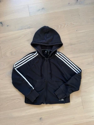 Svart Adidas hoodie med dragkedja - Svart hoodie från Adidas med klassiska vita ränder längs ärmarna och dragkedja framtill. Modellen har huva, två fickor och ribbade muddar. Perfekt för en sportig och avslappnad stil. Materialet är mjukt och bekvämt, troligtvis bomullsblandning.