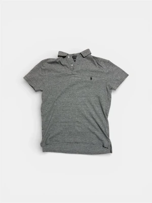 Grå pikétröja från Ralph Lauren - Klassisk grå pikétröja från Ralph Lauren med korta ärmar och krage. Tröjan har två knappar vid halsen och den ikoniska lilla logotypen broderad på bröstet. Tillverkad i mjuk bomull för en bekväm känsla.