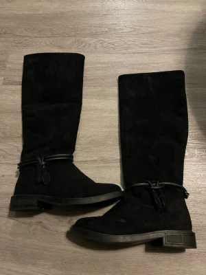 Svarta knähöga mockastövlar - Snygga svarta stövlar i mocka med knähögt skaft och dekorativt band med tofsar runt vristen. Klassisk och stilren design med låg klack och rundad tå. Perfekta för att lyfta din outfit under kallare dagar.