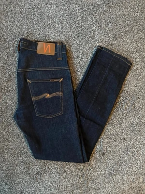 Mörkblå jeans från Nudie - Säljer ett par mörkblå jeans från Nudie Jeans med klassisk femficksmodell och kontrastsömmar. Jeansen har en smal passform och är tillverkade i stretchigt denimtyg för extra komfort. Snygg detalj med Nudie-loggan på bakfickan.