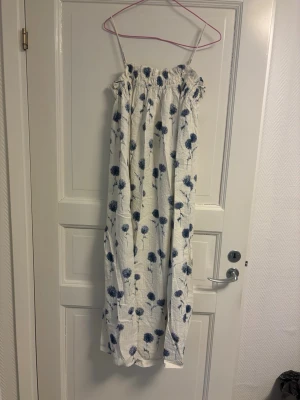 Vit blommig långklänning H&M - Långklänning från H&M i vitt med blått blommönster. Klänningen har tunna axelband och smock upptill för en bekväm passform. Perfekt för varma dagar. Enkel och luftig design som känns somrig och fräsch.