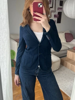 Mörkblå slimfit kavaj med fickor - Snygg mörkblå kavaj i mjukt denim-material med två stora framfickor och klassisk krage. Kavajen har knappstängning och dekorativa knappar vid ärmsluten. Perfekt för en avslappnad men ändå stilren look. Storlek 40 men liten i storleken, passar en storlek 36. 