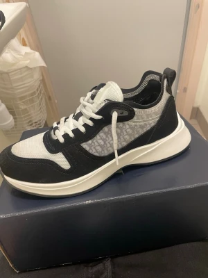 Svarta och vita sneakers från Dior - Snygga sneakers från Dior i svart och vitt med mönstrad mesh på sidan, vita skosnören och tjock vit sula. Skon har en modern design med kontrasterande material och detaljer, samt en pull tab bak för enkel påtagning.