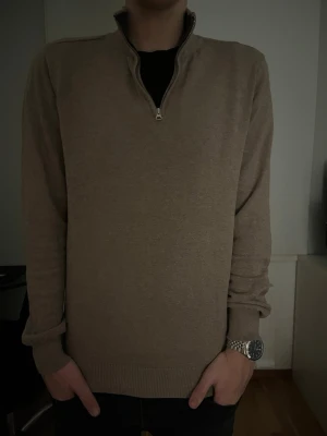 Beige halvzip tröja från John Henric  - Snygg beige tröja med halv dragkedja och ribbade muddar. Tröjan har en klassisk passform och är tillverkad i mjuk bomull, perfekt för lager-på-lager. Enkel och stilren design som funkar till många outfits.