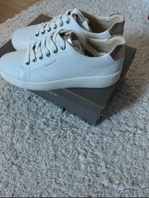 Vita sneakers från Gant med metallicdetalj - Stilrena vita sneakers från Gant med klassisk låg profil och snygga metallicdetaljer vid hälen. Skorna har vita snören, metallfärgade öljetter och är tillverkade i slätt läder. Perfekta för dig som gillar en clean och modern look.