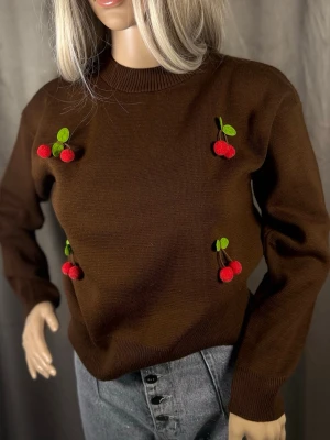 Brun stickad tröja med körsbär - Mysig brun stickad tröja med broderade körsbärsdetaljer i rött och grönt framtill. Tröjan har rund halsringning, långa ärmar och ribbade muddar. Perfekt för dig som gillar lekfulla detaljer och vill sticka ut lite extra.