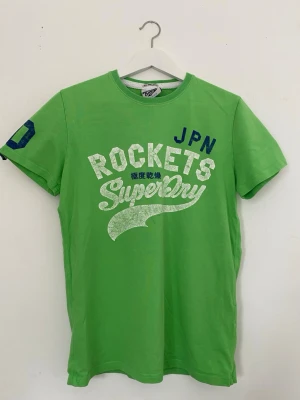 Vintage från Superdry t-shirt med tryck - Snygg grön t-shirt från Superdry med stort vitt och blått tryck på bröstet där det står 'ROCKETS Superdry JPN'. Klassisk rund hals och korta ärmar. Perfekt för dig som gillar sportig och avslappnad stil. Köpt tidigt 2000-tal