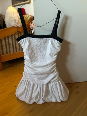 Zara ballongklänning - Säljer en söt vit kort klänning med svarta axelband och kant. Klänningen har en rynkad midja och volang nedtill, vilket ger en feminin siluett. Materialet känns som bomull eller syntet och den är ärmlös med breda axelband.