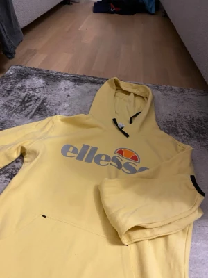Gul Ellesse hoodie med logga - Snygg gul hoodie från Ellesse med stor logga i grått och orange framtill. Klassisk huva med svarta snören och magficka. Perfekt för dig som vill ha en streetwear-look och gillar färg. Mjuk bomullskänsla och avslappnad passform.