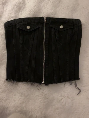 Svart jeanskorsett från Zara - Säljer en svart korsettopp från Zara i jeansmaterial. Toppen är axelbandslös med dragkedja framtill, dekorativa fickor med knappar och rå kant nertill för en edgy look. Perfekt att styla med höga jeans eller kjol. 