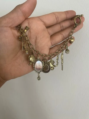 Vintageinspirerat charmarmband i guldfärg - Unikt armband i antikguldfärgad metall med flera olika berlocker som spegel, nyckel, mynt, hjärta och pärldetalj. Kedjan är grov och berlockerna har en cool, retro känsla. Perfekt för dig som gillar statement-accessoarer och detaljer.