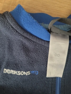 Mörkblå fleecebody Didriksons - Mysig mörkblå fleecebody från Didriksons med blå muddar och dragkedja framtill. Broderad logga på bröstet och mjukt material som passar perfekt för lager-på-lager. Långa ärmar och ben med stretchiga muddar för extra komfort.