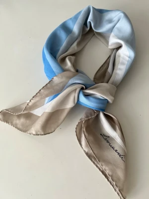 Leonardi silkesscarf i blått och beige - Snygg silkesscarf från Leonardi med breda ränder i ljusblått, beige och vitt. Scarfen har en kvadratisk form och en elegant glansig finish. Perfekt att knyta runt halsen för en stilren look.