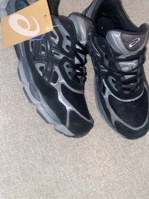 Asics Gel Nyc - Snygga sneakers från Asics i svart och grått med meshpaneler och detaljer i syntet. Skorna har en chunky sula och klassisk snörning. Perfekta för dig som gillar sportig stil och vill ha bekväma skor till vardags.