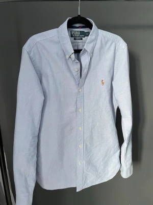 Ljusblå skjorta från Polo Ralph Lauren - Klassisk ljusblå skjorta från Polo Ralph Lauren i slim fit med button-down krage och broderad logga på bröstet. Skjortan har långa ärmar och knäppning framtill. Perfekt för en stilren look.