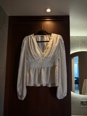 Gina tricot - Vit blus  - Elegant vit blus med djup v-ringning, bred smockad midjedetalj och långa puffärmar med resår vid ärmslut. Materialet är väldigt stretchigt.