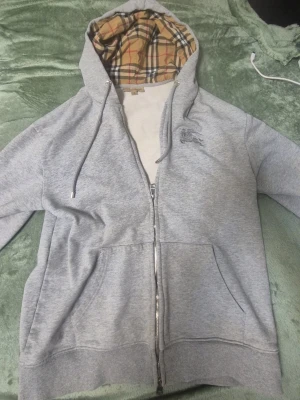 Grå zip-hoodie från Burberry M - Grå hoodie med dragkedja från Burberry. Klassisk huva fodrad med det ikoniska Burberry-rutiga mönstret i beige, svart, rött och vitt. Känguruficka framtill och justerbara snören i huvan. Perfekt för en stilren och avslappnad look.