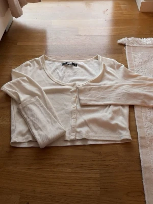 Vit ribbad crop top med knappar - Säljer en vit långärmad crop top med ribbad struktur och knappar framtill. Tröjan har en djup rund halsringning och är tillverkad i ett mjukt, stretchigt material. Perfekt att styla med höga jeans eller kjol.