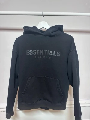 Svart Essentials Fear of God hoodie - Snygg svart hoodie från Essentials Fear of God med stor logga på bröstet. Klassisk känguruficka framtill och huva. Tillverkad i mjukt material som känns skönt mot huden. Perfekt för en avslappnad och trendig streetwear-look.Perfekt för dig som vill ha ett plagg som kombinerar komfort, kvalitet och exklusivt varumärke. Nypris betydligt högre. 📦 Kan skickas snabbt 💬 Hör av dig vid frågor eller om du vill ha fler bilder