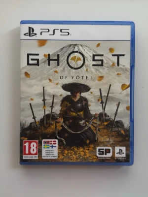 Ghost of Yotei PS5 spel - Ghost of Yotei till PS5 är ett actionäventyrsspel med japanskt tema. Omslaget visar en samuraj med svärd framför ett berg och fallande löv. Spelet kommer i ett blått plastfodral och har åldersmärkning 18+.