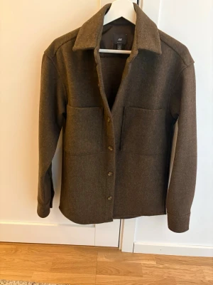 Brun overshirt från H&M - Sitter som en S. Stilren brun overshirt från H&M med klassisk krage och stora bröstfickor. Rak passform och knäppning framtill med matchande knappar. Materialet ser ut att vara en mjuk ullblandning som ger en snygg och avslappnad look. Perfekt lager på lager-plagg.