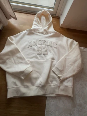 Vit hoodie med text Angelic 92 - Säljer en oversized vit hoodie med huva och stor magficka. På bröstet står det 'ANGELIC 92' i upphöjd text. Hoodien har ribbade muddar vid ärmslut och nederkant. Perfekt för en chill och avslappnad stil. Materialet känns mjukt och bekvämt.