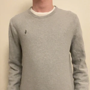 Ljusgrå tröja från Polo Ralph Lauren - Säljer en stilren ljusgrå tröja från Polo Ralph Lauren med rund halsringning och diskret broderad logga på bröstet. Tröjan har en klassisk passform och är perfekt för en clean och enkel look.