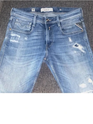 Blå slitna jeans från Replay - Säljer ett par blå jeans från Replay med slitningar och distressed detaljer. Klassisk femficksmodell med snygg tvätt och Replay-logga på fickan. Perfekta för en avslappnad och trendig look.