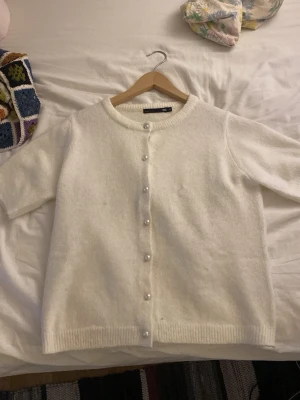 Vit kofta med pärlknappar från lager157 - Söt vit kofta från lager med korta ärmar och rund halsringning. Koftan har dekorativa pärlknappar framtill och är stickad i ett mjukt material. Perfekt att slänga över en topp för en clean och stilren look. Aldrig använd