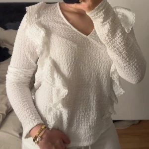Vit blus med volang från Vila - Säljer en superfin vit blus från Vila med v-ringning och långa ärmar. Blusen har en strukturerad, lite bubblig yta och dekorativa volanger som går från axlarna ner längs framsidan. Perfekt för dig som gillar romantiska detaljer och en feminin look.