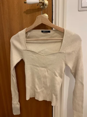 Beige ribbad topp från Gina Tricot - Trendig beige långärmad topp från Gina Tricot med ribbad struktur och fyrkantig urringning. Figurnära passform och mjukt stretchigt material som sitter snyggt på kroppen. Perfekt att styla med jeans eller kjol.