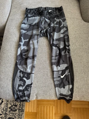 Nike Pro camo tights  - Säljer ett par Nike Pro tights med grått och svart camo-mönster. De har en bred resår i midjan med Nike Pro-logga och en vit Nike Swoosh på benet. Materialet är stretchigt och andas, perfekt för träning och sport.