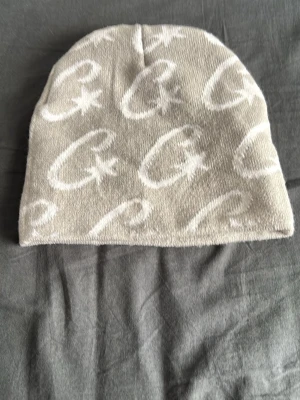 Cortez mössa  - Säljer en beige mössa med vita grafiska bokstavsmönster och stjärnor. Mössan är mjuk och stretchig, perfekt för kyliga dagar. Materialet känns som bomull eller en bomullsblandning. Enkel och stilren design som passar till det mesta.