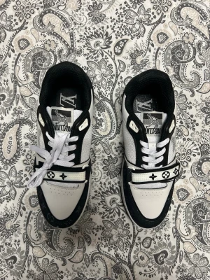 L-V svartvita sneakers - Snygga svartvita sneakers från L-V med klassiska monogramdetaljer och signatur på sidan. Skorna har vita snören, svart sula och kontrasterande paneler i läder och textil. Perfekta för dig som vill sticka ut med exklusiv streetstyle.