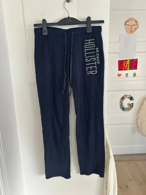 Mörkblå mjukisbyxor från Hollister - Snygga mörkblå mjukisbyxor från Hollister med vit logga och texten 'SO. CALIF' längs ena benet. Byxorna har dragsko i midjan och raka ben. Perfekta för chill dagar eller när du vill ha det extra bekvämt. Förekommer några fläckar som ej syns jätte mycket 