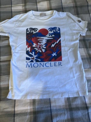 Vit Moncler t-shirt med tryck - Snygg vit t-shirt från Moncler med färgstarkt tryck framtill i blått, rött och vitt som föreställer en solnedgång och naturmotiv. Klassisk passform och rund halsringning. Perfekt för dig som vill sticka ut med en exklusiv look.
