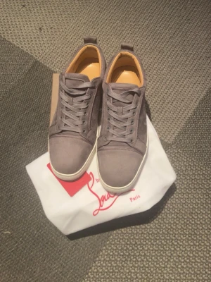 Gråa mockasneakers från Louboutin - Säljer ett par stilrena gråa sneakers från Christian Louboutin i mocka med vita sulor och klassisk röd detalj på undersidan, clean design som passar till det mesta. Storlek 42 men kan även passa 43