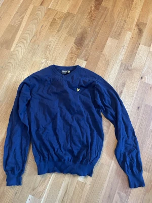 Mörkblå stickad tröja Lyle & Scott - Mörkblå stickad tröja från Lyle & Scott med rund halsringning och klassisk gul logga på bröstet. Tröjan har långa ärmar och ribbade muddar vid ärmslut och nederkant. Perfekt för dig som gillar stilren och enkel design.