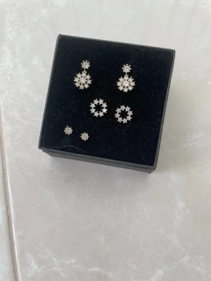 Set med blommiga örhängen i silver - Säljer ett set med tre par örhängen i silverfärg, alla med blommiga former och gnistrande stenar. Två par är större och cirkelformade, ett par är små och diskreta. Perfekta för att mixa och matcha till olika looks.