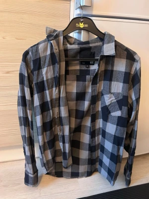 Rutig skjorta i bomull  - Säljer en klassisk rutig skjorta i svart och grått. Skjortan har lång ärm, bröstficka och knappar framtill. Perfekt för en avslappnad och trendig look. Tillverkad i mjuk bomull för extra komfort.