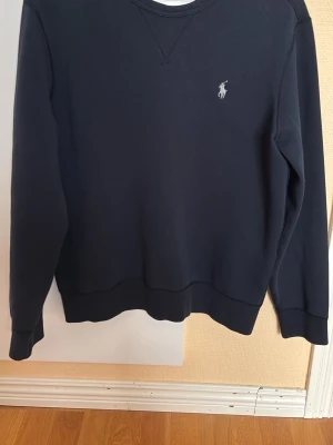 Mörkblå sweatshirt från Ralph Lauren - Klassisk mörkblå sweatshirt från Ralph Lauren med rund halsringning och den ikoniska vita logon broderad på bröstet. Tröjan har ribbade muddar vid ärmslut och nederkant, samt en stilren och enkel design som passar till det mesta.