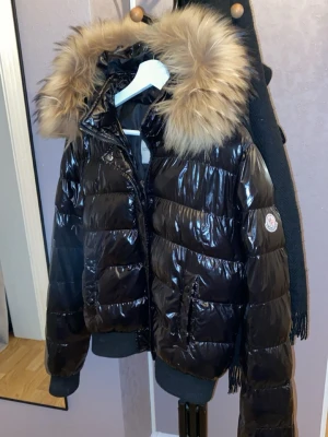 Svart glansig dunjacka Moncler - Snygg svart glansig dunjacka från Moncler med stor fluffig fuskpälskrage på huvan. Jackan har ribbade muddar, två framfickor och klassisk Moncler-logga på ärmen. Perfekt för kalla vinterdagar och riktigt trendig look. Har ingen aning om den är äkta eller inte, använt bara en gång och den känns som ny även fast den är köpt för länge sedan. Inga skador eller smuts på den. 