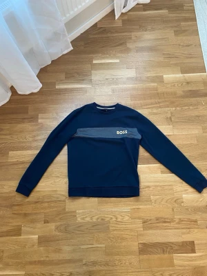 BOSS Tröja  - Snygg mörkblå sweatshirt från BOSS med en grå panel över bröstet och vit BOSS-logga. Tröjan har rund halsringning och långa ärmar. Perfekt för en clean och stilren look.